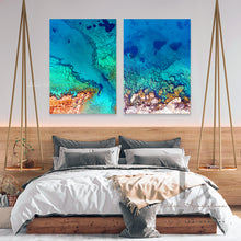 A large coastal-inspired wall art set on canvas the wall, over bed, in boho bedroom decor setting. The room features  white walls, a bed surrounded with gray pillows, plans, and a decorative boho accents. The coastal Aerial wall art canvas set features,  turquoise waters, rock, pebbles, making an relaxing, cosy and calming atmosphere.