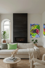 A modern living room featuring a large black fireplace with a built-in fire, a cream-colored sofa with green and gray pillows, and two colorful abstract canvas paintings hanging on the walls.