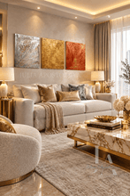 A luxury modern living room featuring a beige sofa with gold and brown accent pillows, a marble coffee table, and three vertical abstract paintings in gold, copper, and silver tones on the textured beige wall.