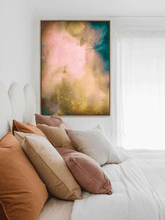 A framed abstract wall art print with soft pink, teal, and gold tones hanging above a bed with white bedding and multiple blush, tan, and rust pillows in a bright bedroom.