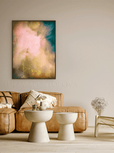 A vertical framed abstract canvas print with soft pink, teal, and gold brushstrokes, displayed on a beige wall above a brown leather sofa in a modern living room setting.
