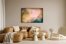 An abstract canvas print with soft pink, gold, and teal tones hanging on a beige wall above a brown leather sectional sofa in a modern living room setting.