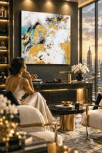 A woman in a cream satin gown sits in a luxurious modern living room, facing a large abstract canvas painting with gold, white, and teal brushstrokes. The room features a dark wood console table, gold leaf accents, and a large window overlooking a city skyline at sunset.