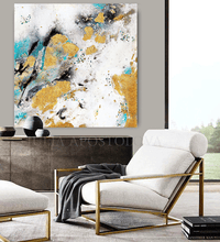 An abstract canvas wall art with gold leaf, teal, black, and white brushstrokes, displayed in a modern living room setting above a white armchair with a gold frame.