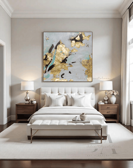 Abstract painting with gold leaf accents hanging above a neatly made white bed in a neutral-toned bedroom with wooden bedside tables and white lamps, accompanied by a large white velvet coffee table. The room has large windows allowing natural light , and the decor includes decorative vase with pale rose flowers and a light beige-colored rug on the floor. The overall aesthetic is cozy and contemporary.