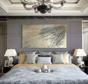 A gold wabi sabi painting with 3d relief gold and silver textures covering the entire canvas. The painting is displayed on the wall of a luxury bedroom with a large window overlooking a natural landscape. The room features a gray bed frame and nightstand, silver decorative pillows, and a coffee service tray , on the bed creating a warm and inviting atmosphere