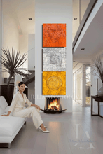 A woman in a white outfit sits on a white sofa in a modern living room, facing a tall vertical triptych wall art piece with three textured panels in copper, silver, and gold tones above a fireplace.