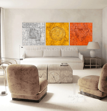 A modern living room featuring a three-panel canvas wall art set by Julia ApostoloVA. The artwork consists of three textured square panels in gray, gold, and copper, depicting abstract circular and organic patterns.