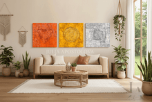 A styled living room featuring a three-piece canvas wall art set by artist Julia Apostoloa. The set consists of three textured square canvases in vibrant orange copper, golden yellow, and cool silver gray, hung in a row above a beige sofa.