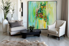 A large framed abstract painting with vibrant green, yellow, and brown brushstrokes hangs on a light gray wall in a modern living room setting with a beige sofa, white armchair, black coffee table, and a fluffy beige rug.