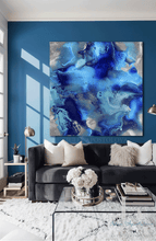 An abstract canvas wall art print featuring swirling shades of luminous blue, navy, and light teal with silver metallic accents, displayed in a modern living room setting above a dark gray sofa.
