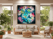 A large framed abstract canvas print with vibrant splashes of blue, green, pink, and purple hangs on a beige wall in a modern, sunlit living room with a rattan sofa, chair, and plants.