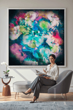 A woman sits in a gray lounge chair in a modern living room, holding a magazine and cup, with a large colorful abstract canvas painting displayed on the wall behind her.