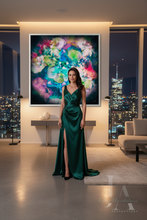 A woman stands in a modern living room wearing a floor-length emerald green satin gown with a thigh-high slit. Behind her is a large framed abstract floral painting, with city windows and a sofa visible in the background.