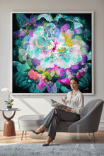 A woman sits in a gray lounge chair holding a magazine in a modern living room, with a large framed abstract floral painting hanging on the wall behind her. The artwork features a vibrant mix of teal, purple, pink, blue, and green tones.