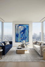 A large framed abstract painting with vibrant blue, navy, and silver brushstrokes hangs on a white wall in a modern, sunlit living room with a blue sofa, a light gray sofa, and a city view through tall windows.