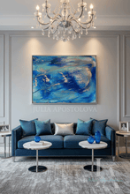 A modern living room featuring a large abstract blue painting on a gray wall above a deep blue sofa. The room is styled with a crystal chandelier, marble coffee tables, blue glass vases, and a textured gray rug.