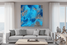 A large square abstract canvas print with vibrant blue, teal, and silver swirls, displayed on a gray wall above a modern light gray sofa in a bright, contemporary living room.
