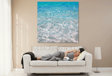 Woman lying on a couch reading a book with a large ocean painting above her in a living room.