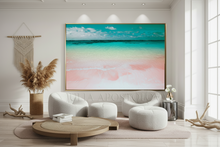 A large framed wall art print depicting a tropical beach scene with turquoise water, white sand, and pink shoreline, displayed in a modern living room setting with white tufted chairs and a round coffee table.