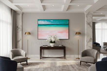 A framed beach art print with turquoise ocean and pink sand hangs on a light gray wall in a modern, elegant living room with gray armchairs, a console table, and gold floor lamps.