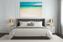 A large square canvas wall art print depicting a tropical beach scene with turquoise ocean water and a sandy shore, displayed in a modern bedroom above a bed with white linens.