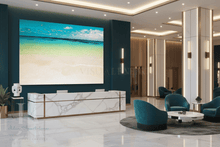 A large wall-mounted beach scene print displayed in a modern lobby reception area with marble reception desk, teal chairs, and warm-toned interior columns.
