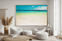 A large framed beach art print hangs on a white wall in a modern, sunlit living room. The artwork by artist Julia Apostolova, depicts a tropical ocean scene with turquoise water, pale sand, and a blue sky.
