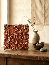 A square copper-colored textured wall art plaque displayed on a rustic wooden table, styled with a ceramic vase, dried branches, and a small bowl in a warm, neutral-toned interior setting.