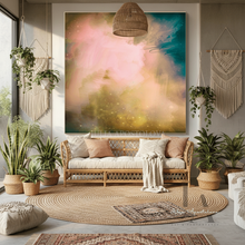 A large abstract wall art print featuring pastel pink, teal, and gold nebula-like colors, displayed above a rattan sofa in a styled boho living room with plants, macrame wall hangings, and woven rugs.