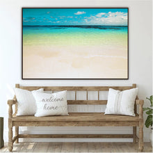 A large framed beach art print hangs on a white wall above a wooden bench with white pillows. The artwork depicts a tropical shoreline with turquoise water, pale sand, and a blue sky.