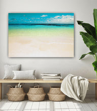 A large rectangular wall art print showing a tropical beach with turquoise water, pale sand, and a blue sky, displayed above a bench in a styled interior with pillows, books, baskets, and a plant.