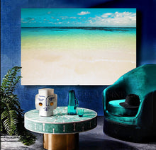 A large rectangular wall art print showing a tropical beach with turquoise water, pale sand, and blue sky, displayed in a styled interior above a round table and next to a teal armchair.