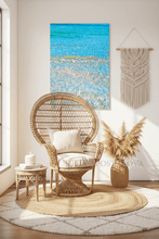 Wicker chair with white cushion in a room with a large abstract painting on the wall of turquoise clear waters.