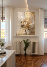 An abstract gold and white painting, displayed in a gold frame on a white wall above a shelf in a modern, sunlit room with white curtains and wood flooring.