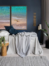 A large coastal-inspired minimalist beach wall art set on the wall, over bed, in boho bedroom decor setting. The room futures navy blue walls, a gray woolen carpet, bed surrounded with pillows, plans, a low wooden coffee table and a decorative boho accents.