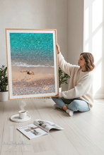 A woman sits on a light wood floor holding a large framed beach print by Artist Julia Apostolova. The print shows turquoise water, a sandy shore, and small rocks, with a light wood frame.