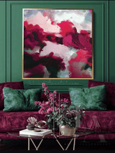 Abstract canvas wall art with bold magenta, pink, sage green, and white brushstrokes in a gold frame, displayed above a deep green velvet sofa with matching pillows in a styled living room setting.e green