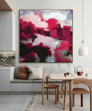 A large framed abstract canvas print featuring bold magenta, pink, sage green, and white brushstrokes, displayed on a light gray wall above a modern dining area with wooden chairs and a table.