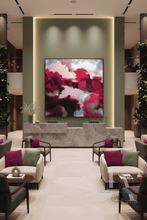 A large abstract canvas painting with vibrant magenta, pink, sage green, and black brushstrokes hangs on a modern beige wall in a luxury lobby. The painting is displayed above a marble reception desk, flanked by symmetrical cream armchairs with magenta and sage green pillows, and framed by tall green plants.