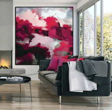 A large abstract canvas wall art print featuring vibrant magenta, pink, red, and white brushstrokes with black accents, displayed in a modern living room setting above a dark gray sofa.