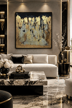 An abstract gold and black painting with textured metallic gold brushstrokes and gold leaf on a gray background, hangs above a white sofa in a modern luxury living room with marble furniture and warm lighting.