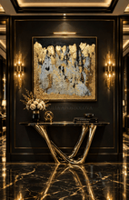 An abstract gold and black painting with textured metallic gold brushstrokes and gold leaf on a gray background, displayed in a luxurious black entryway with gold trim, a polished marble floor, and a metallic console table below.