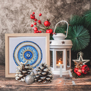 A white lantern with a lit candle inside, surrounded by pine branches , red berries , pine cones , and a framed original glitter artwork with a colorful mandala design on a wooden table