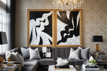 
Two black and white abstract paintings with organic shapes displayed above a grey sofa in a room with gold accents.
