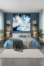 A large square abstract canvas print with blue, white, and black brushstrokes hangs on a dark blue accent wall above a neatly made bed in a modern, styled bedroom.