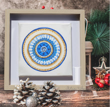 A framed original painting with circle mandala-style glitter Greek evil eye, surrounded by pine branches , red berries , pine cones , and Christmas ornaments , on a wooden table. Ideal art gift for Christmas, birthday, and any occasion.