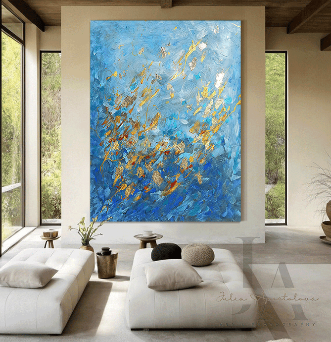 Abstract artwork with blue and gold tones , featuring swirling patterns and textures . The background is a modern interior with large windows showing greenery outside . There are two white sofas with cushions in the foreground and a small plant on a table 