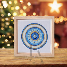 A framed original greek evil wall art in circular mandala design with intricate patterns and colors, including shades of blue , white , and gold , set against a blurred background with Christmas decorations. Will be perfect elegant gift for someone special.