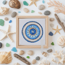 A square framed originalart featuring a concentric blue and gold circular design with a small clear rhinestone at the center, surrounded by beach-themed decorative items like shells, stones, coral, driftwood, and starfish on a white background. A cursive watermark reading 'Julia Anastasia' is visible at the bottom.